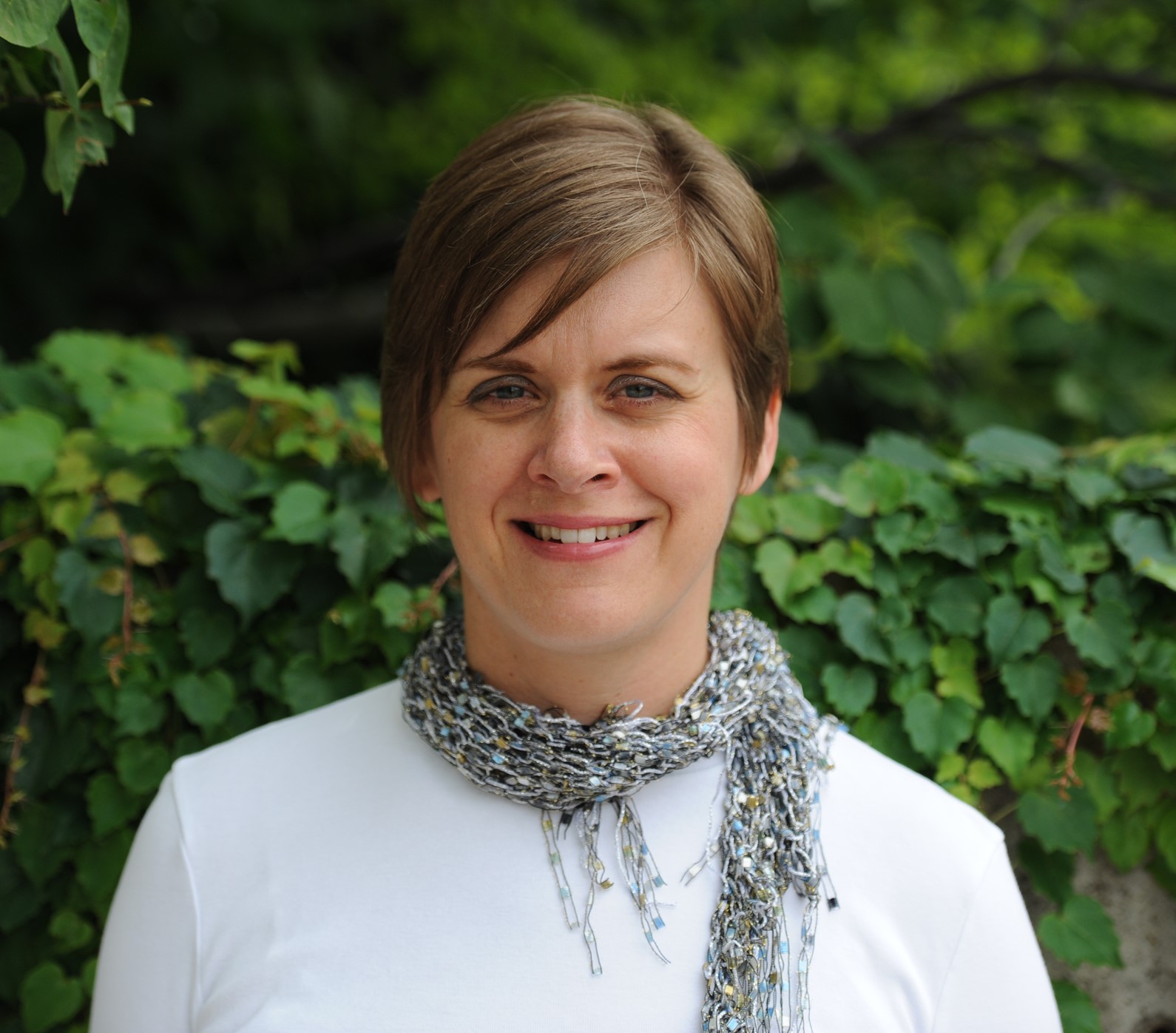 A headshot with short, light brown hair and light colored skin, wearing a white shirt and a grey scarf.